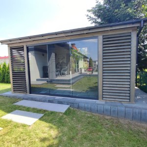 A modern wooden structure with large glass windows and decorative slats, set in a grassy area. The interior features seating and is designed for outdoor living.